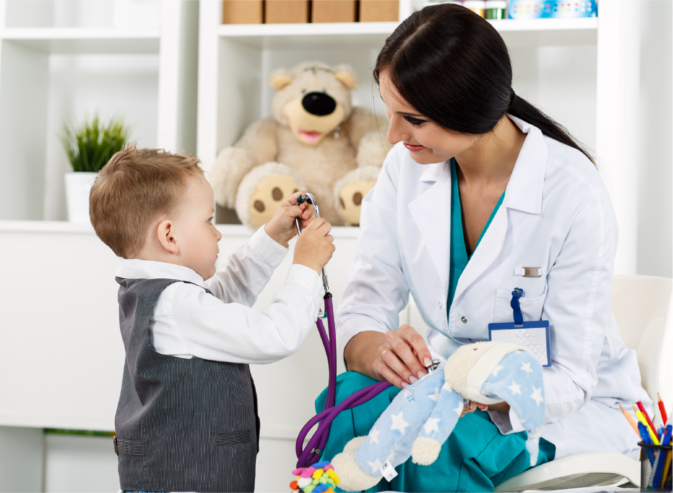 Pediatric doctor in white lab coat letting young toddler boy hold stethoscope during child wellness visit
